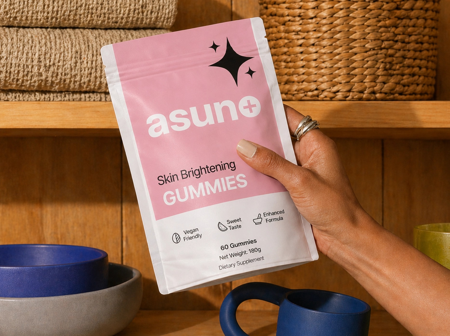 Hand holding pink ASUNO Skin Brightening Gummies 60 vegan friendly sweet taste dietary supplement package against wooden shelf with bowls and baskets